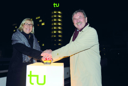 Eine Frau und ein Mann drücken auf einen Knopf. Im Hintergrund leuchtet das TU Logo.