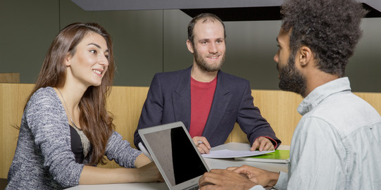 Eine Frau und ein Mann sitzen gegenüber von einem Mann welcher auf seinem Laptop tippt