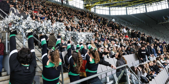 Eine Stadiontribüne voller junger Menschen, unten stehen Cheerleader in schwarz-grünen Kostümen und silbernen Pompons.