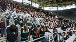 Eine Stadiontribüne voller junger Menschen, unten stehen Cheerleader in schwarz-grünen Kostümen und silbernen Pompons.