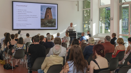 Foto: Eine Person spricht vor einem sitzenden Publikum bei einer Veranstaltung in einem Ausstellungsraum.