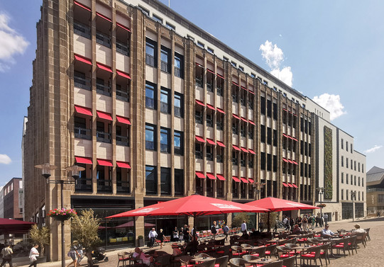 Kaufhaus mit Außenterrasse. Die Fenster haben rote Blenden, im Vordergrund stehen rote Sonnenschirme.