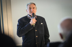 Foto: Eine Person hält ein Mikrofon und spricht während einer Veranstaltung vor Publikum.