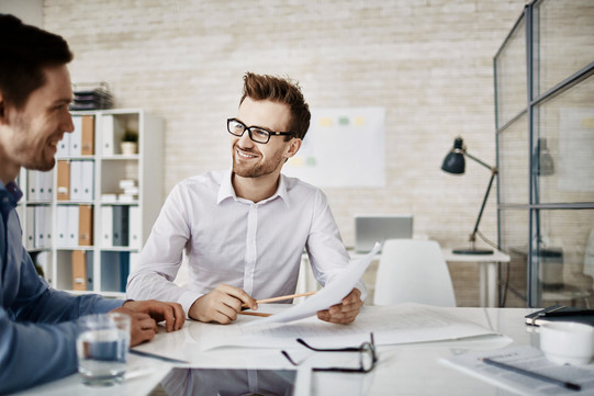 Two men in business shirts sitting at a desk with documents, smiling and talking to each other