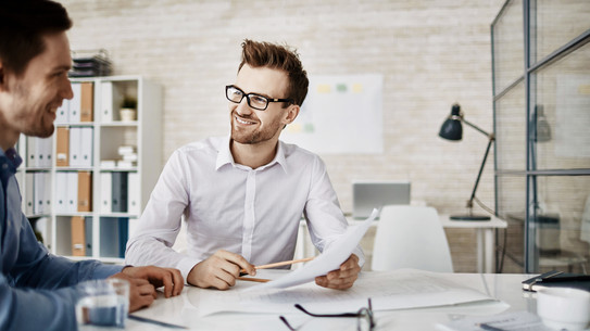 Zwei Männer unterhalten sich. Vor ihnen liegen Dokumente auf einem Tisch ausgebreitet.