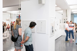 In the center of the picture is a white wall fixture with pictures hanging on it. Two people are looking at them. To the left and right of the wall, you can see a large exhibition space with other people interested in art.