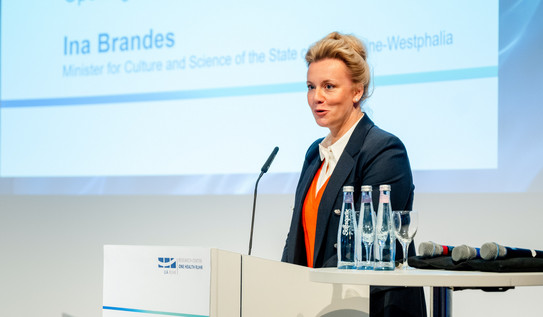 Photo: Science Minister Ina Brandes stands at a lectern at the opening of the One Health Ruhr Research Center. Brandes wears a blonde updo, a black blazer and a red blouse.