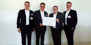Gruppenfoto: Vier Personen stehen nebeneinander, zwei von ihnen halten in der Mitte gemeinsam einen Förderbescheid in den Händen.