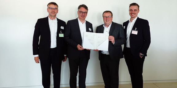 Gruppenfoto: Vier Personen stehen nebeneinander, zwei von ihnen halten in der Mitte gemeinsam einen Förderbescheid in den Händen.