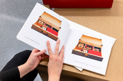 Ausstellung „Laufende Beobachtungen“ In einer Ausstellung halten zwei Hände ein Buch. Im Buch sind zwei Boxen mit geöffneten Schubladen zu sehen.