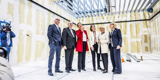 Foto: Eine Gruppe von sechs Personen steht in einem unfertigen Raum mit freiliegenden Wänden und Deckenbalken und blickt gemeinsam nach oben.