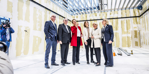 Foto: Eine Gruppe von sechs Personen steht in einem unfertigen Raum mit freiliegenden Wänden und Deckenbalken und blickt gemeinsam nach oben.