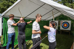 Foto: Zwei Personen üben Bogenschießen an einem Stand auf dem Sommerfest. Zwei andere Personen erklären ihnen die richtige Haltung.