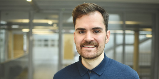 Porträt von JProf. Tim Heubeck: JProf. Heubeck hat braune Haare und trägt ein blaues Oberteil.