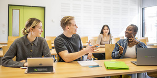 Studenten sitzen in einem Seminarraum an Tischen. In der vorderen Reihe sprechen ein Student und eine Studentin lächelnd miteinander, während die anderen ihnen zuhören.