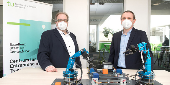 Zwei Männer mit Anzug und Mund-Nase-Bedeckung stehen vor einem Tisch mit zwei Robotern und zwei Produktionsstraßen.