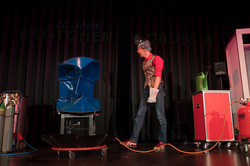 A man in dark trousers and a red shirt walks across a stage. Around him are various experimental set-ups.