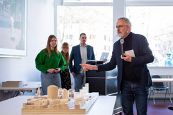 Im Vordergrund des Bildes ist ein Architekturmodell aus Holz zu sehen, welches auf einem Tisch steht. Daneben steht ein Mann im Anzug und hält ein Mikrophon in der Hand. Hinter im stehen drei junge erwachsene Personen und schauen in Richtung des Mannes. Im Hintergrund ist eine große helle Fensterfront zu sehen.