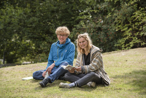 A blond student with a blue jacket and a blonde student with a brown jacket are sitting in a meadow.
