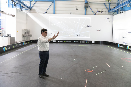 Ein Mann, Hubertus Heil, steht mit erhobenen Händen und einer VR-Brille auf dem Kopf in einem großen Raum, in dem viele Drohnen herumfliegen.