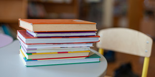 Auf dem Bild sieht man einen Bücherstapel auf einem Tisch liegen. Neben dem Tisch steht ein Stuhl.