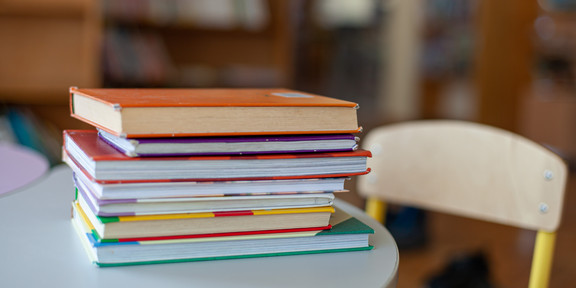 Auf dem Bild sieht man einen Bücherstapel auf einem Tisch liegen. Neben dem Tisch steht ein Stuhl.