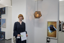 Eine Frau steht vor einer weißen Wand neben einem Kunstprojekt einer Kunstausstellung und hält eine Urkunde in den Händen.