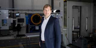 Dr. Till Clausmeyer Dr. Till Clausmeyer im dunkelblauen Anzug mit kurzen hellbraunen Haaren. Im Hintergrund stehen Maschinen in der Experimentierhalle der Fakultät Maschinenbau.