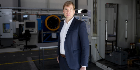 Dr. Till Clausmeyer im dunkelblauen Anzug mit kurzen hellbraunen Haaren. Im Hintergrund stehen Maschinen in der Experimentierhalle der Fakultät Maschinenbau.