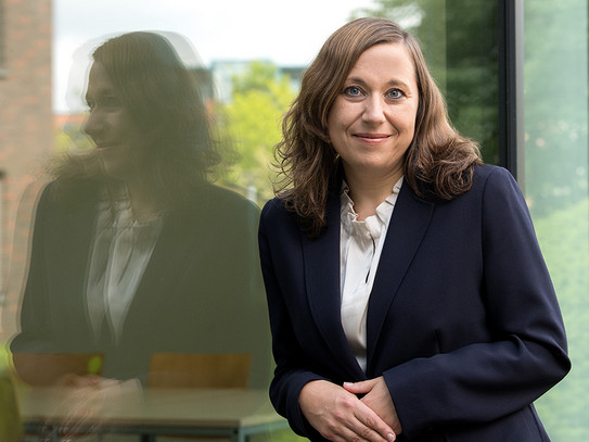 Porträt von Prof. Nadja Dörflinger: Prof. Dörflinger trägt einen dunklen Blazer und lehnt an einer gläsernen Fassade.