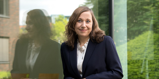 Porträt von Prof. Nadja Dörflinger: Prof. Dörflinger trägt einen dunklen Blazer und lehnt an einer gläsernen Fassade.