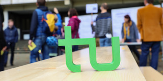 A small green TU logo stands on a wooden table. People walk by in the background.