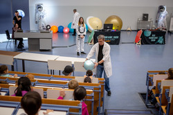 Ein Mann steht in einem vollbesetzten Hörsaal und gibt einen Wasserball, der einen Planeten symbolisieren soll, an im Publikum sitzende Kinder weiter.