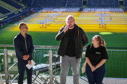 Auf dem Foto stehen zwei Männer und eine Frau auf der Tribüne. Der Mann in der Mitte hält ein Mikrofon in der Hand und spricht in Richtung Publikum.  