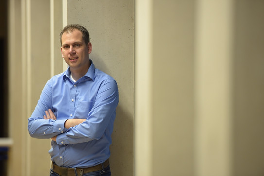 Portrait photo of Daniel Neider leaning against a concrete wall in a blue shirt, arms crossed and smiling slightly.
