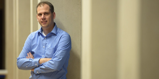 Portrait photo of Daniel Neider leaning against a concrete wall in a blue shirt, arms crossed and smiling slightly.