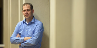 Portraitfoto von Daniel Neider, der in einem blauen Hemd an einer Betonwand lehnt, die Arme verschränkt hat und leicht lächelt.