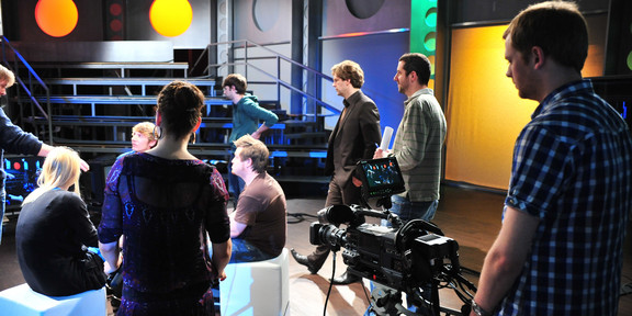 Studio NRWision Personen sitzen im Studio. ein Mann schaut sich das Geschehen durch eine Kamera an