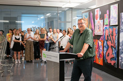 Im Zentrum des Bildes steht Prof. Gerhard Schembecker an einem Pult. Hinter ihm befindet sich eine schwarze Wand mit Kunstwerken von Studierenden sowie Kunstinteressierten.