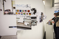 Foto: Im Vordergrund sieht man verschiedene Kunstwerke. Im Hintergrund stehen zwei Frauen, die sich ein Kunstwerk genauer anschauen.