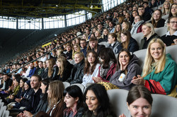 Eine Tribüne voller Studierender, die sich freuen und teilweise in die Kamera lächeln.