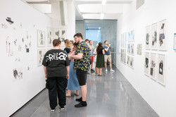 Auf dem Bild ist ein langer Gang zu sehen. An den weißen Wänden links und rechts hängen verschiedene Kunstwerke. Im Gang stehen Menschen, welche sich die Kunstwerke angucken.