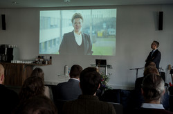 Im unteren Teil des Bildes sind die Hinterköpfe eines Publikums zu sehen, die auf eine Leinwand im Zentrum des Bildes gerichtet sind. Auf der Leinwand ist Ina Brandes zu sehen, eine Ministerin aus NRW.