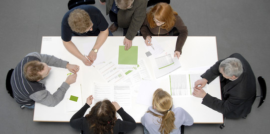 Viewed from above: Seven people sitting around a white table full of documents.