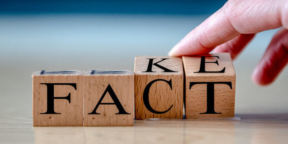 On a table, four wooden cubes with letters that form the word "FACT" are placed next to each other. A hand tilts the last two cubes, on which the letters "KE" can be seen. Thus, both the words "FACT" and "FAKE" are legible.
