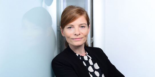 Porträtfoto von Prof. Fanziska Wallner: Prof. Wallner steht vor einem spiegelnden hellen Hintergrund und trägt einen dunklen Blazer.