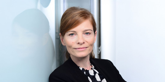 Porträtfoto von Prof. Fanziska Wallner: Prof. Wallner steht vor einem spiegelnden hellen Hintergrund und trägt einen dunklen Blazer.