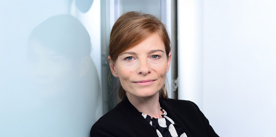 Portrait photo of Prof. Franziska Wallner: Prof. Wallner stands in front of a reflective light background and wears a dark blazer.