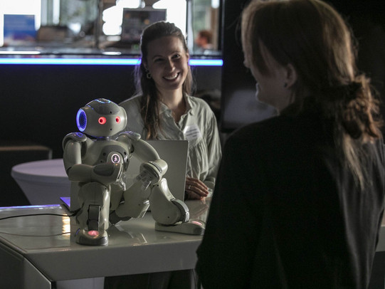 Foto: Ein Roboter sitzt auf einem Tisch, während eine Person davor und eine weitere dahinter steht.