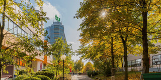 Rechts und links Bäume in herbstlichen rot-brauen Farben und ein TU Dortmund Gebäude mit Logo in der Mitte.